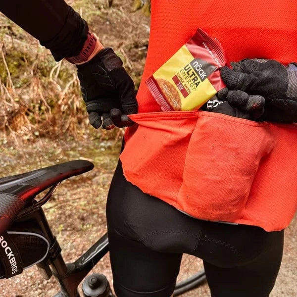 Person in red jacket with a naak ultra energy waffle in a pocket, standing next to a bicycle on a natural background.