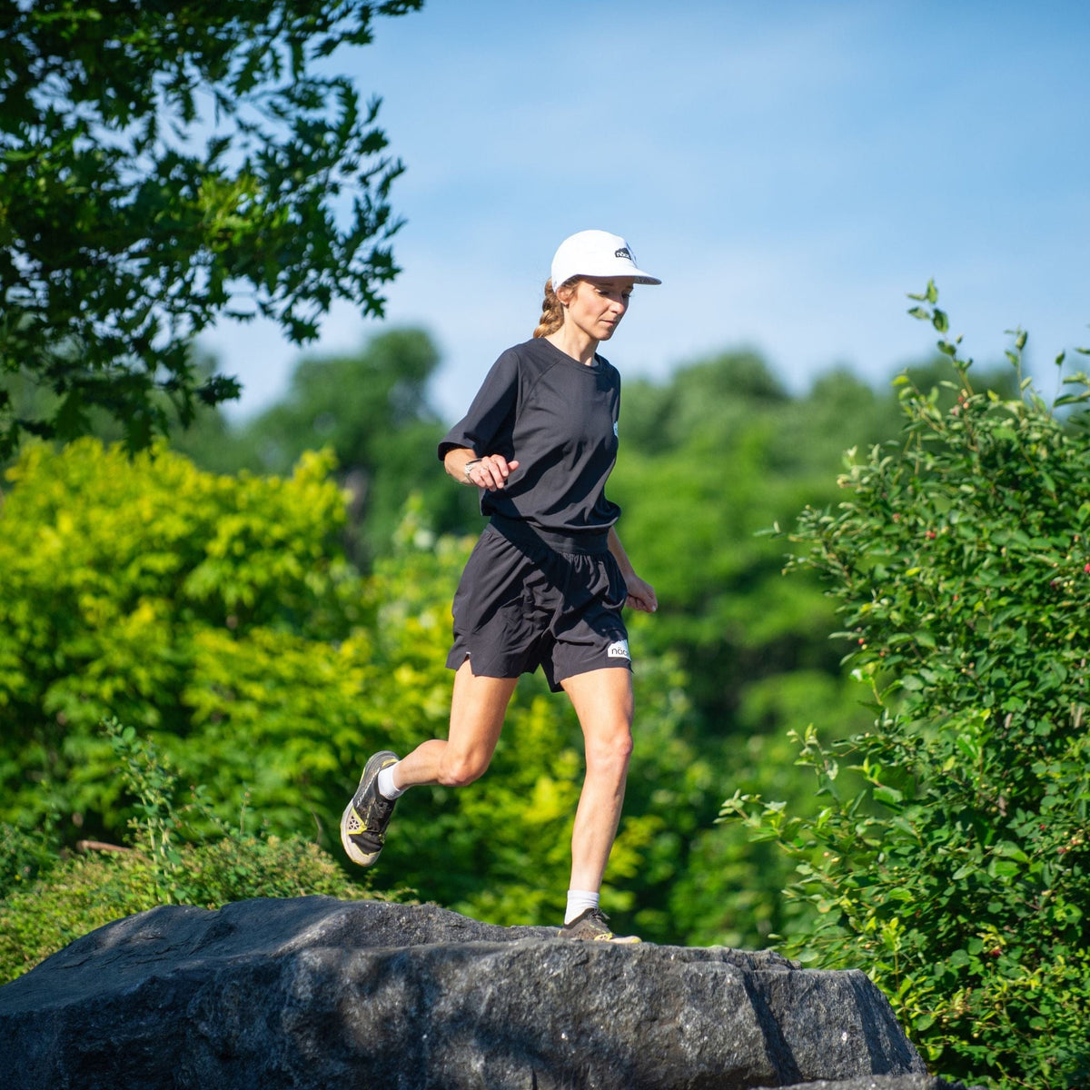 woman athlete runs in naak apparel, black praise t shirt and black praise shorts, with white cap