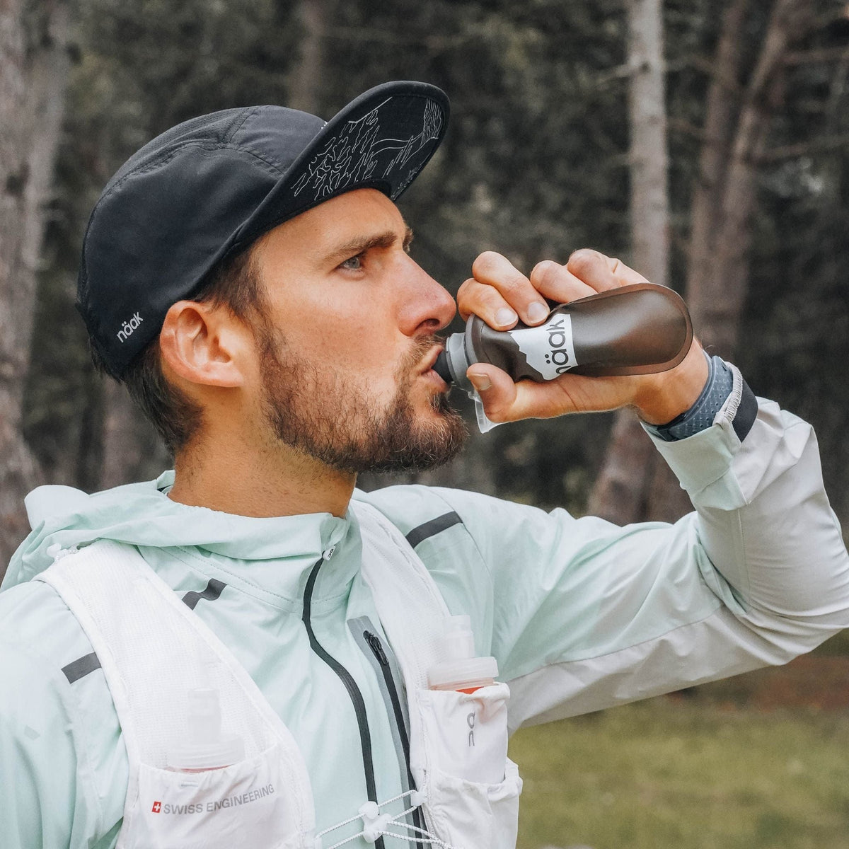 Man drinking from the naak soft flask outdoors with trees in the background