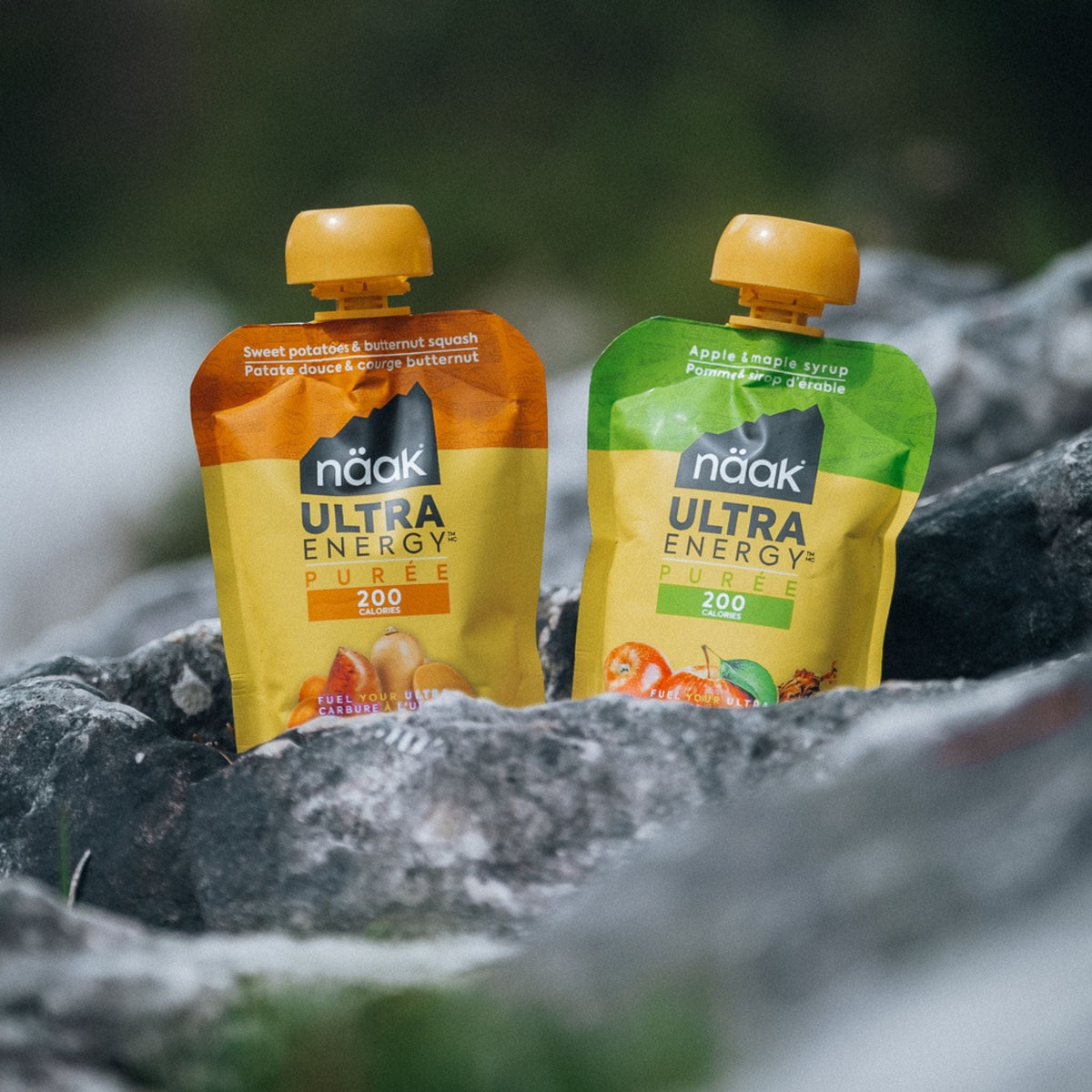 Two naak Ultra Energy Purees packets on a rocky surface with a blurred natural background.