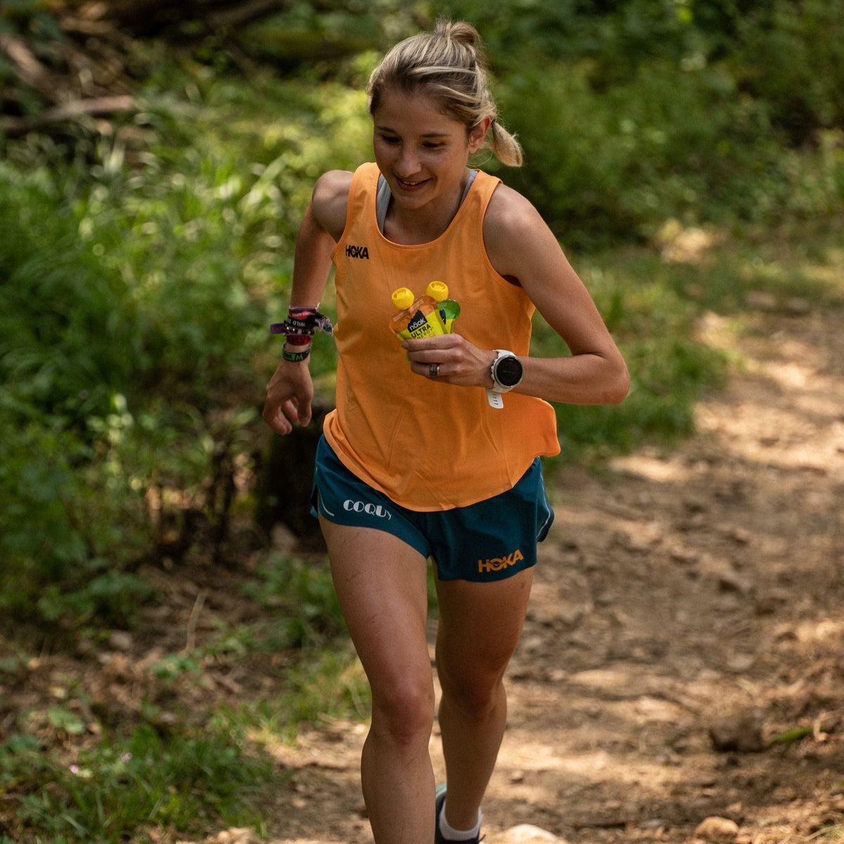 Woman running on a trail wearing an orange tank top and blue shorts holding two naak ultra energy purees sweet potatoe butternut squash 
